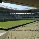 Vista ampla de um estádio de futebol vazio, com campo gramado e arquibancadas sob a luz do dia.
