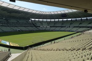Vista ampla de um estádio de futebol vazio, com campo gramado e arquibancadas sob a luz do dia.