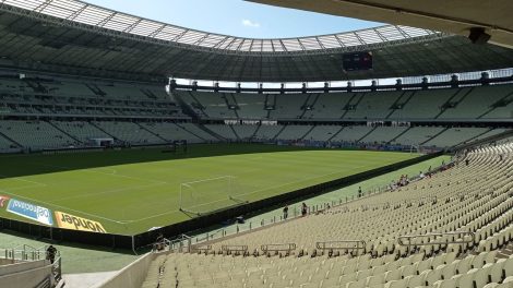 Vista ampla de um estádio de futebol vazio, com campo gramado e arquibancadas sob a luz do dia.