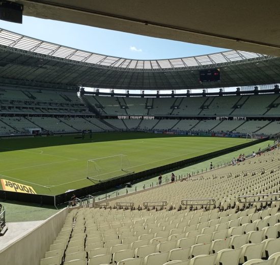 Vista ampla de um estádio de futebol vazio, com campo gramado e arquibancadas sob a luz do dia.