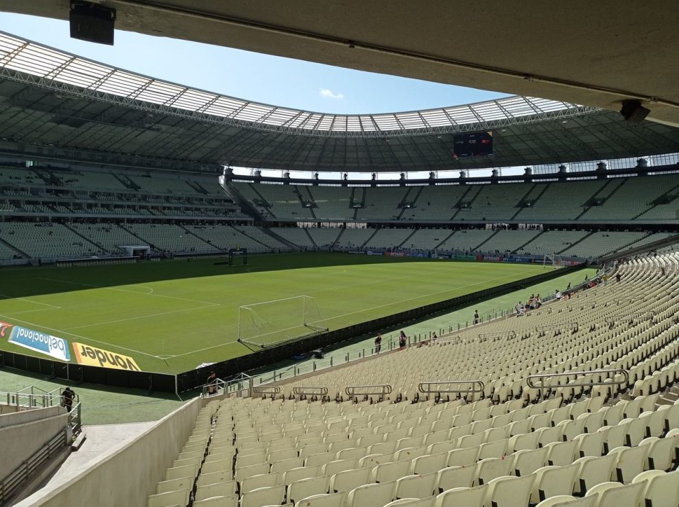 Vista ampla de um estádio de futebol vazio, com campo gramado e arquibancadas sob a luz do dia.