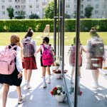 Cinco estudantes com mochilas caminham em direção à entrada da escola em um dia ensolarado.