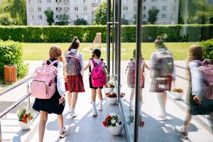 Cinco estudantes com mochilas caminham em direção à entrada da escola em um dia ensolarado.