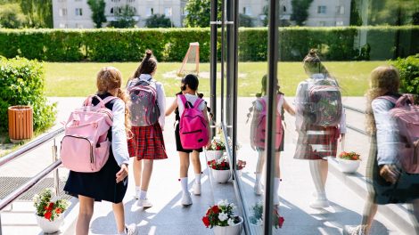 Cinco estudantes com mochilas caminham em direção à entrada da escola em um dia ensolarado.