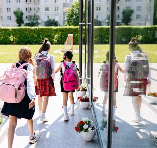 Cinco estudantes com mochilas caminham em direção à entrada da escola em um dia ensolarado.