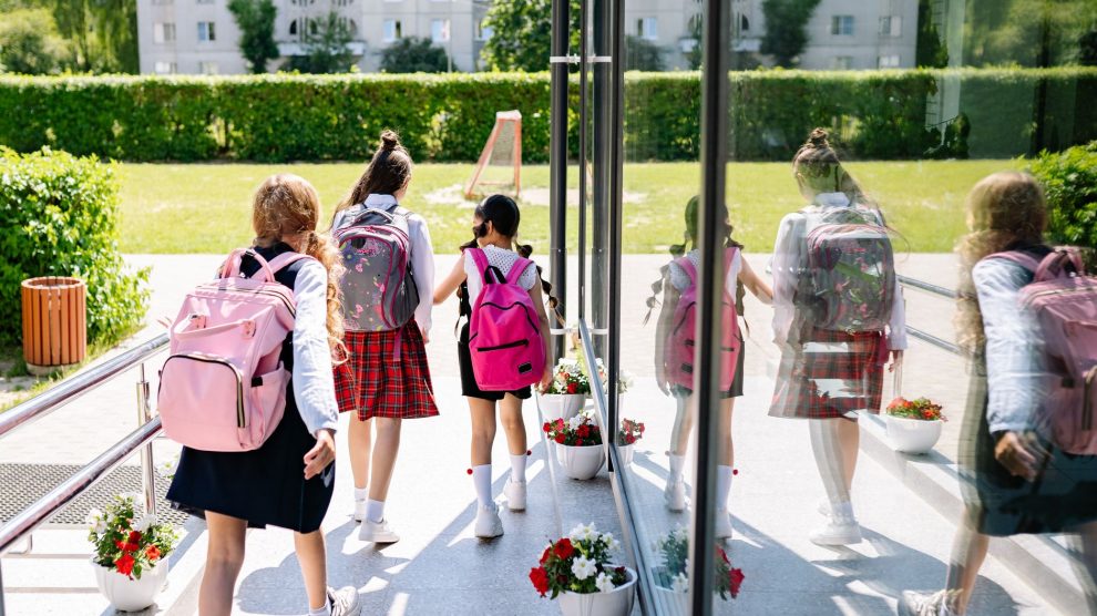 Cinco estudantes com mochilas caminham em direção à entrada da escola em um dia ensolarado.