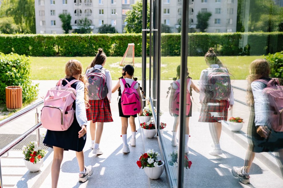 Cinco estudantes com mochilas caminham em direção à entrada da escola em um dia ensolarado.